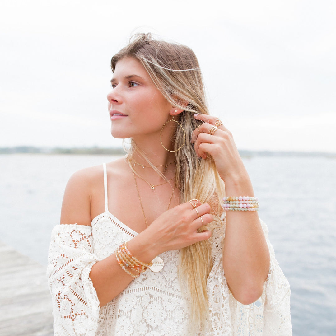 Woman wearing Olive Branch Hoop earrings, layered necklaces and bracelets, standing by the beach in a lace boho dress. The hand-carved gold hoops symbolize peace and resilience, perfect for adding a natural, serene touch to any outfit.