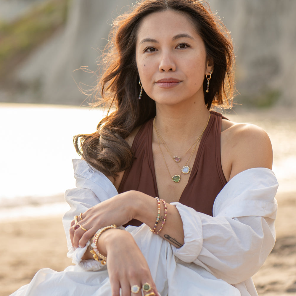 Woman wearing Aurora Fluorite Gemstone Hoop Earrings by Blooming Lotus Jewelry. The delicate fluorite gemstone beads dangle gracefully, adding a subtle pop of color that complements her layered gemstone necklaces and stacked bracelets. She exudes a relaxed, bohemian elegance with a soft beach backdrop, creating a harmonious blend of nature-inspired style.