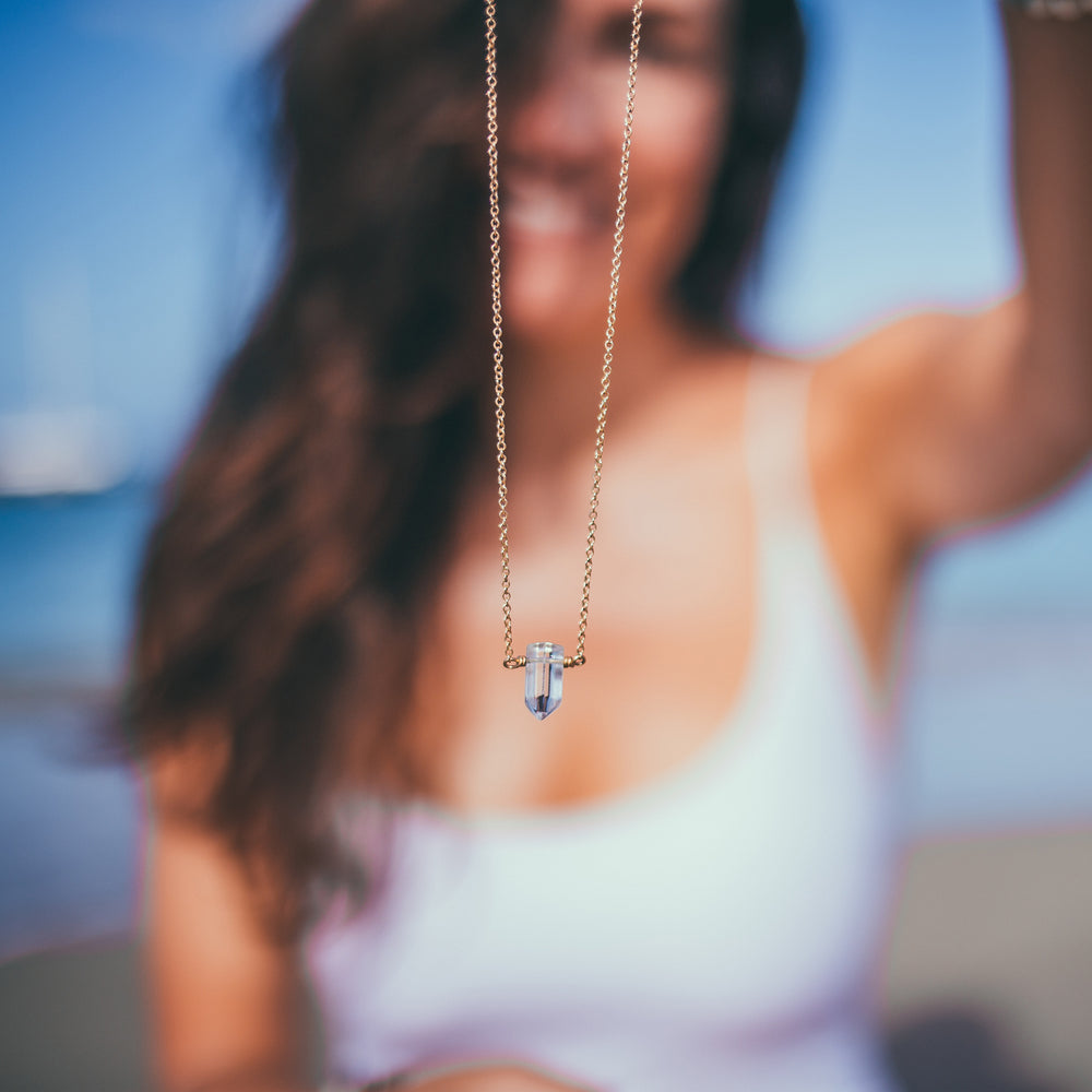 Close-up of a woman holding a gold necklace with a clear crystal pendant, set against a blurred beach background, showcasing a boho-chic and nature-inspired aesthetic.