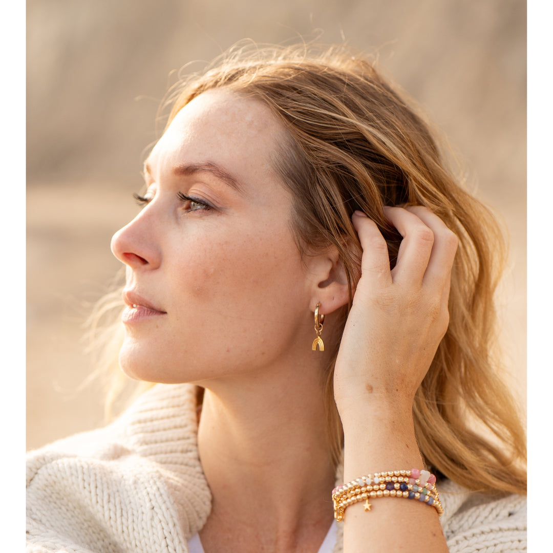 Close-up of a woman wearing Chasing Rainbows Gold Hoop Earrings by Blooming Lotus Jewelry. She gently touches her hair, showing the rainbow charm that dangles from the hoop. Her natural makeup and soft waves create a serene, beach-inspired look, complemented by a stack of beaded bracelets on her wrist, reflecting boho and nature-inspired style.