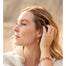 Close-up of a woman wearing Chasing Rainbows Gold Hoop Earrings by Blooming Lotus Jewelry. She gently touches her hair, showing the rainbow charm that dangles from the hoop. Her natural makeup and soft waves create a serene, beach-inspired look, complemented by a stack of beaded bracelets on her wrist, reflecting boho and nature-inspired style.