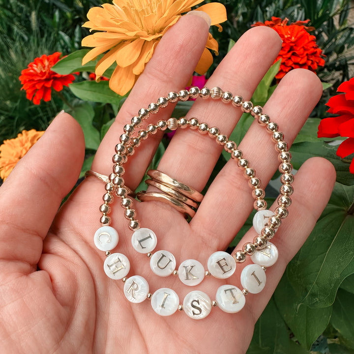 Hand holding two gold beaded bracelets with mother-of-pearl letter beads spelling "LUKE" and "CHRISTIAN," accented with small gold spacer beads. Set against a backdrop of vibrant orange and red flowers.