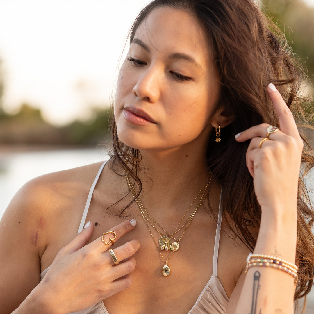 Woman wearing Eye of Protection Hoop Earrings by Blooming Lotus Jewelry. The gold hoops feature a small, intricate evil eye charm, symbolizing protection. She complements the earrings with layered gold necklaces, rings, and bracelets, creating a refined, boho-inspired look that exudes a sense of tranquility by the water.