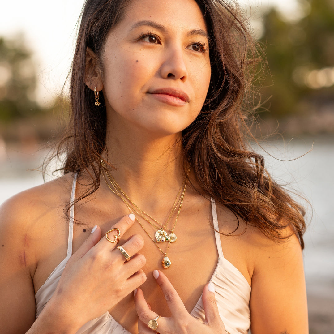 Woman wearing Eye of Protection Hoop Earrings by Blooming Lotus Jewelry. The delicate evil eye charms on the gold hoops symbolize protection and complement her layered gold necklaces and rings. Bathed in soft sunset light by the water, she exudes a calm, boho-inspired elegance, creating a serene, beachy aesthetic.