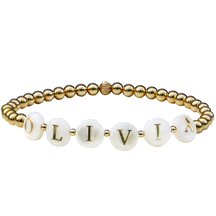 Gold beaded stretch word or name bracelet with mother-of-pearl letter beads spelling "OLIVIA," accented with small gold spacer beads. A customizable bracelet that can be personalized with names or words.