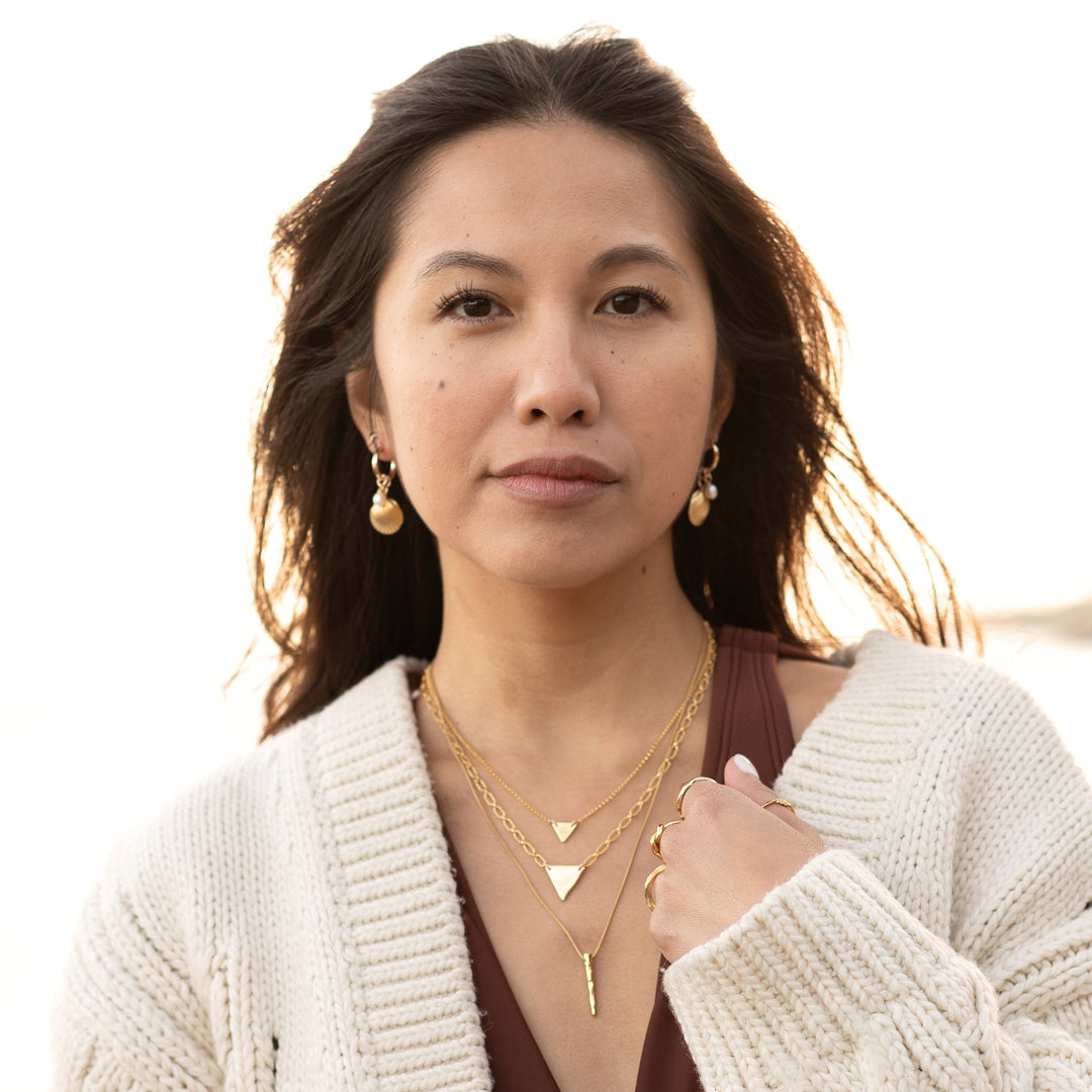 Woman wearing Golden Tide Shell Pearl Hoop Earrings by Blooming Lotus Jewelry. The gold hoops feature textured shell charms and freshwater pearls, adding a subtle elegance to her beach-inspired look. Paired with layered gold necklaces and rings, her style is effortlessly chic, capturing a warm, coastal vibe in natural light.