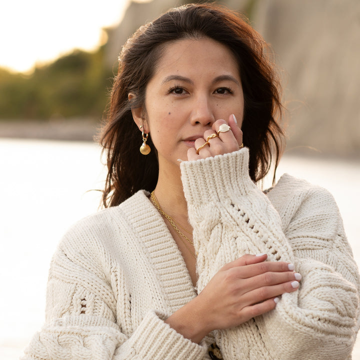 Woman wearing Golden Tide Shell Pearl Hoop Earrings by Blooming Lotus Jewelry. The earrings feature textured gold shell charms paired with delicate freshwater pearls, adding a coastal elegance to her cozy, natural look. She holds a chunky knit sweater close, enhancing the warm, beach-inspired aesthetic against a soft, serene backdrop.