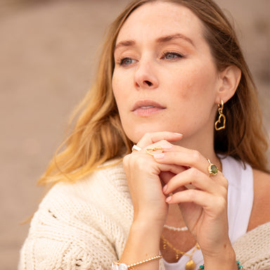 Woman wearing Heart of Gold Hoop Earrings by Blooming Lotus Jewelry. The open heart-shaped charm hangs gracefully from her ears, adding a touch of elegance to her soft, contemplative expression. She rests her hands near her face, showing off gold rings and a delicate bracelet, enhancing her natural, boho-inspired look.