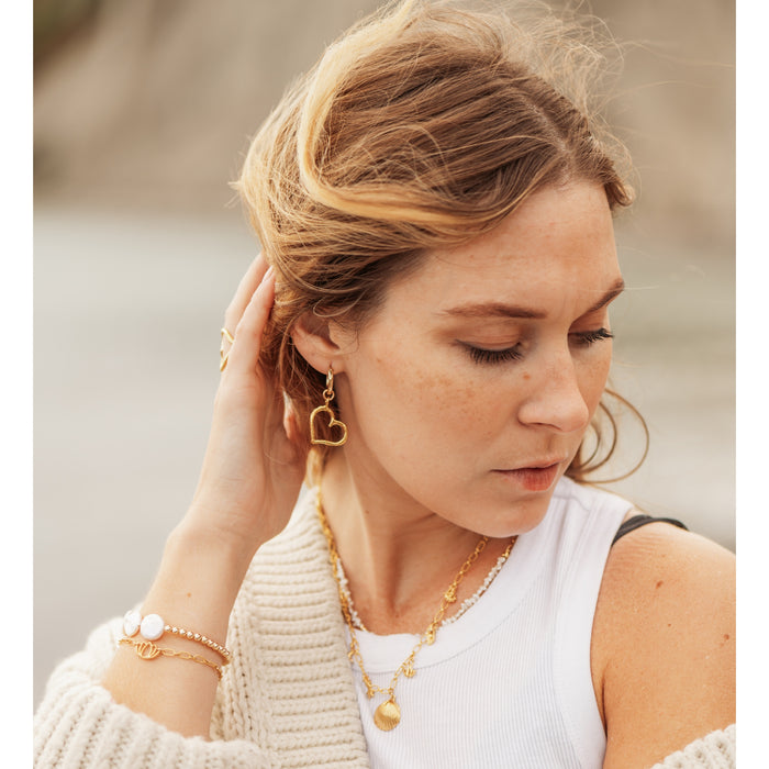 Close-up of a woman wearing Heart of Gold Hoop Earrings by Blooming Lotus Jewelry. The open heart-shaped charm dangles elegantly from the gold hoop, exuding a romantic and timeless feel. She lightly touches her hair, with layered gold necklaces and a bracelet stack adding to her relaxed, boho-inspired beach style.