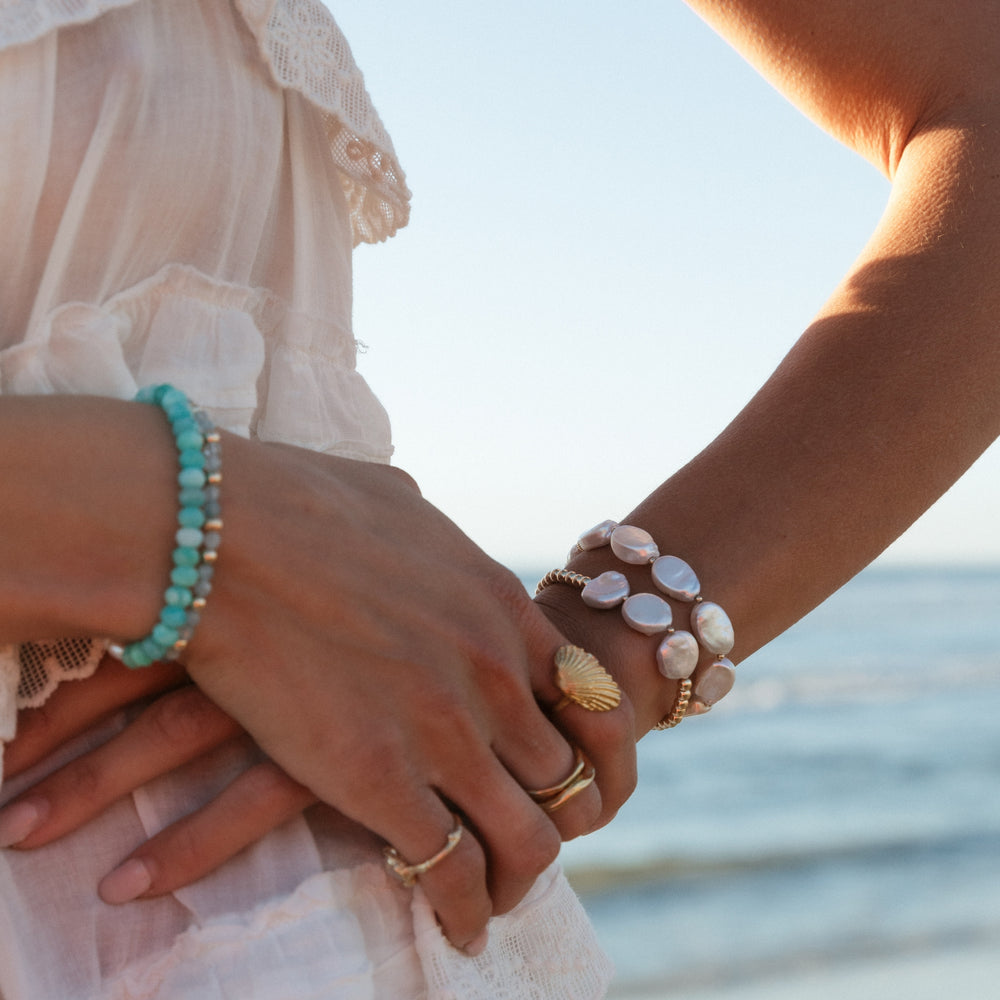 Close-up of a woman’s hands adorned with layered gold rings, a turquoise beaded bracelet, and stacked pearl bracelets, set against a serene beach backdrop with ocean waves.