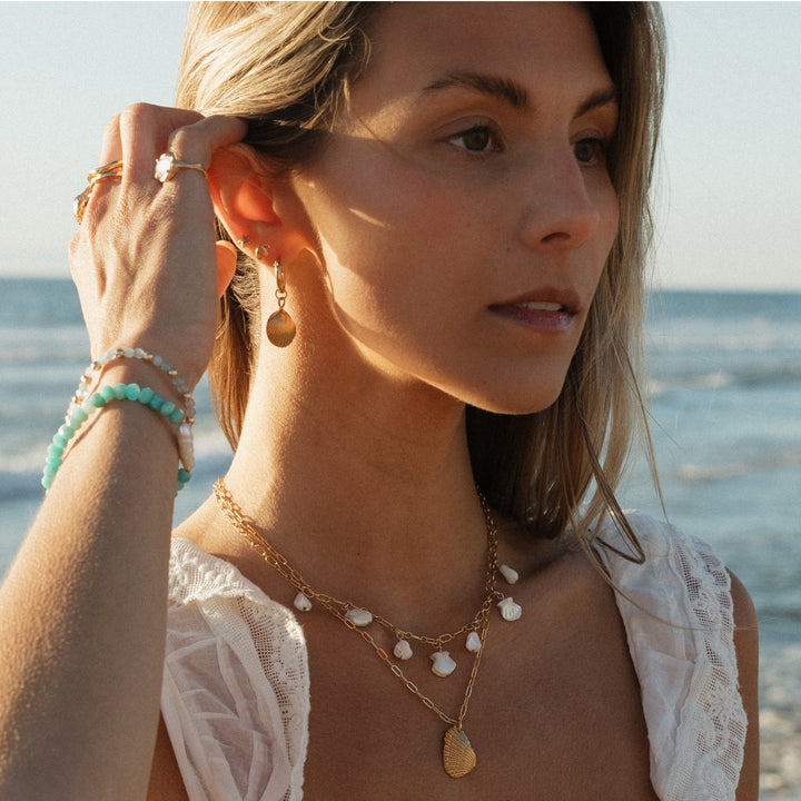 Sunlit close-up of a woman wearing layered gold shell necklaces, matching shell drop earrings, and ocean-inspired bracelets, with the beach and waves in the background, capturing effortless coastal elegance.