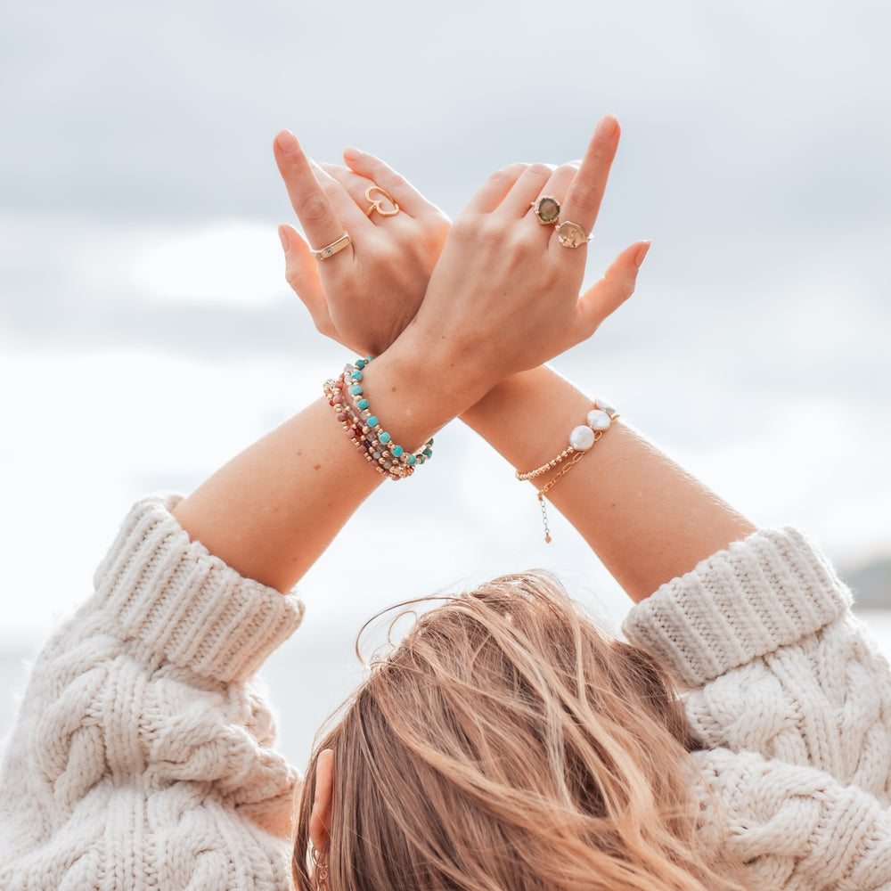 Close-up of a woman’s hands raised against a cloudy sky, adorned with layered gemstone bracelets, including a turquoise Laguna bracelet, gold beaded bracelets, and pearl accents, along with gold rings, capturing a free-spirited boho vibe.