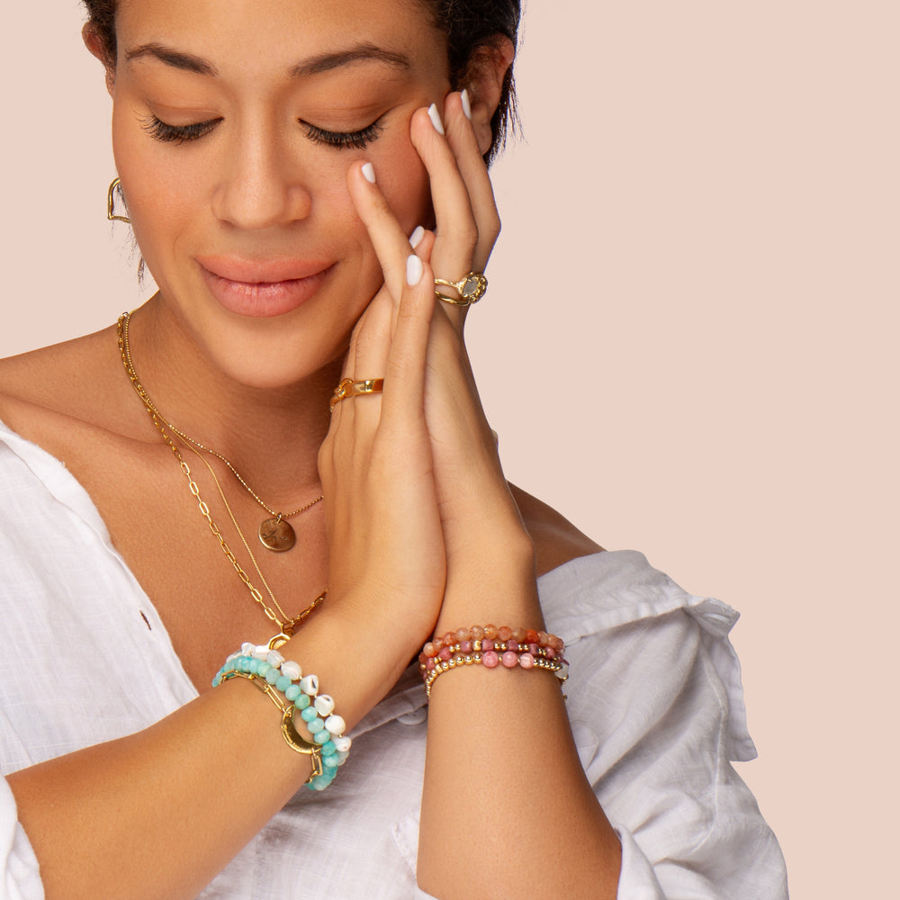 Close-up of a woman with a serene expression, wearing layered gold necklaces, rings, and stacked gemstone bracelets, including a Rhodonite 'Let Love Flow' bracelet and an ocean-inspired Amazonite and Mother of Pearl bracelet, exuding a soft, boho-chic style.