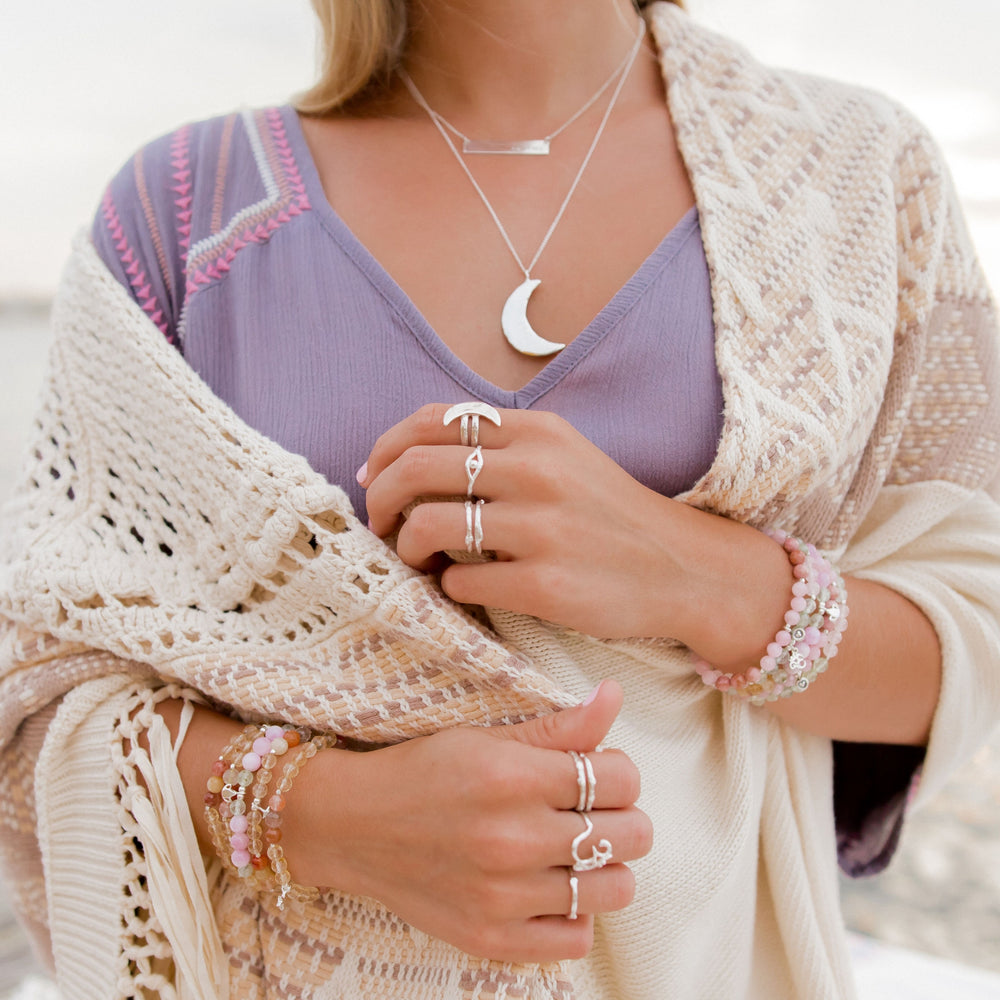 Close-up of a woman wearing layered celestial jewelry, including the Luna Crescent Moon Ring and other silver stacking rings. She is styled with a crescent moon necklace, delicate bracelets with pink gemstones, and a cozy boho knit shawl over a lavender top, creating a dreamy and boho look.