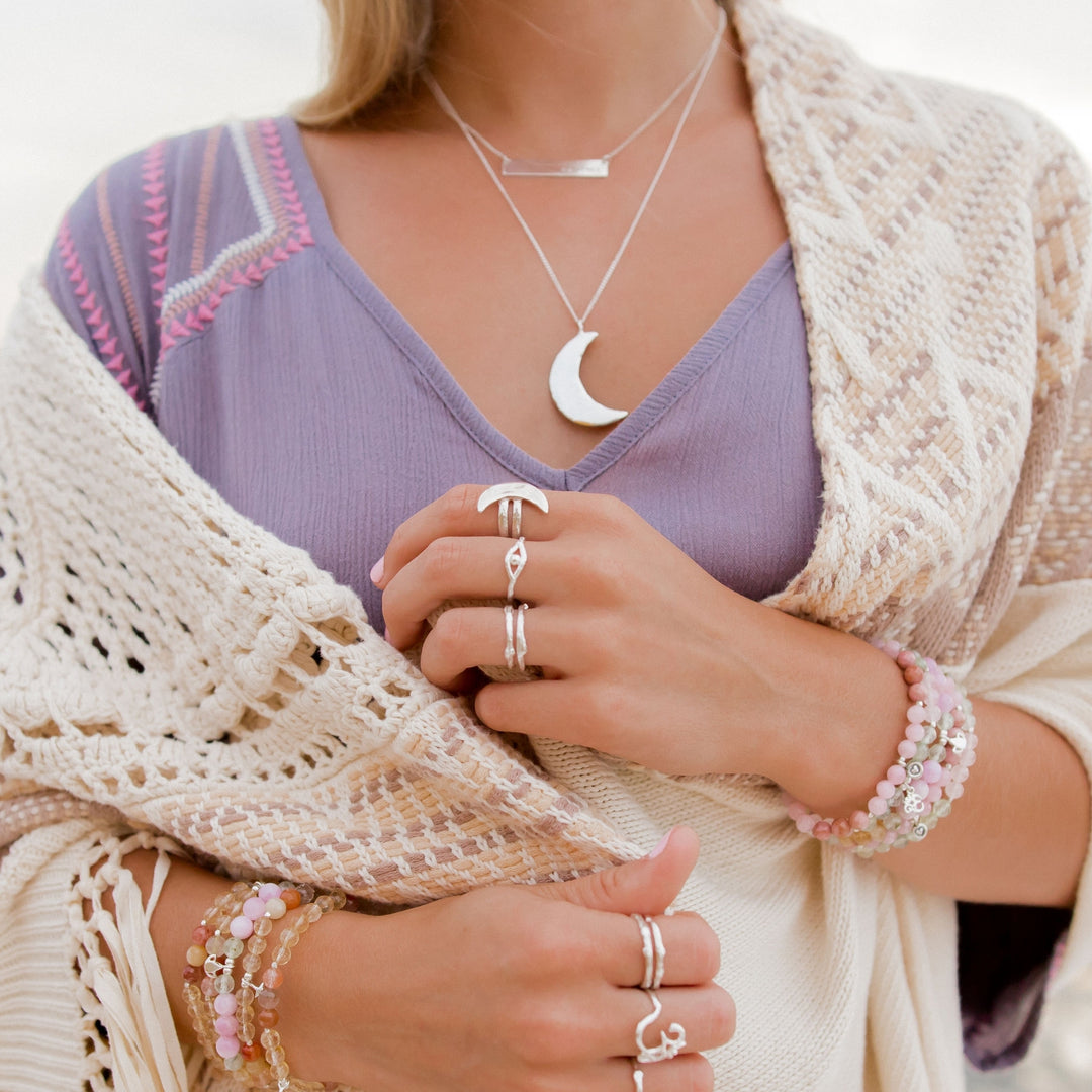 Close-up of a woman wearing layered silver necklaces, including the Luna Crescent Moon Necklace, paired with silver rings featuring celestial and boho designs. She is dressed in a lavender top with embroidered details, wrapped in a cozy crochet shawl, and accessorized with pastel gemstone bracelets, creating a soft, dreamy boho look.