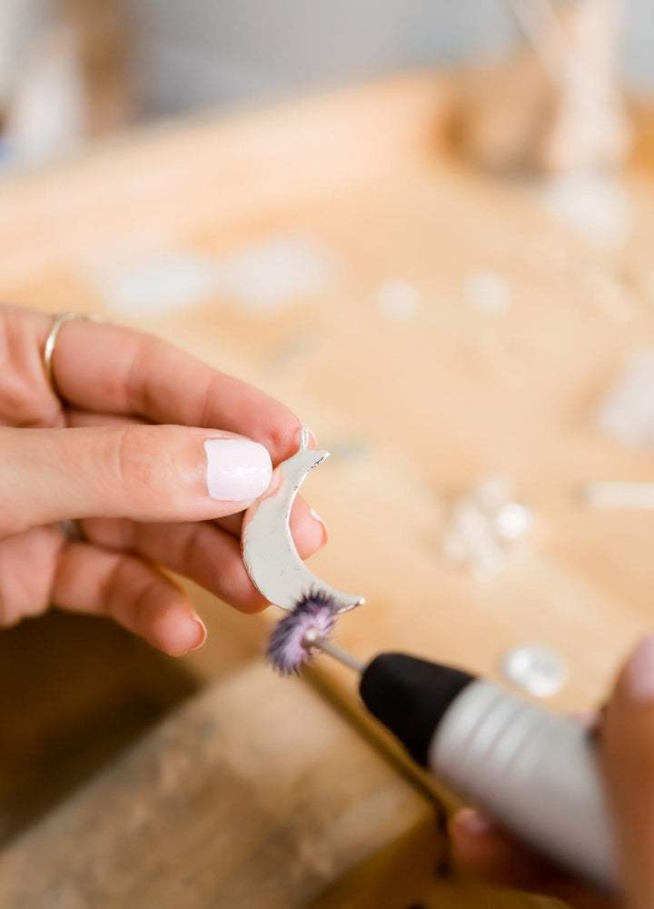 Close-up of hands polishing a crescent moon-shaped silver pendant with a rotary tool, showcasing the craftsmanship involved in creating the Luna Crescent Moon Necklace. The pendant is held delicately against a wooden workbench background, emphasizing the handmade process.