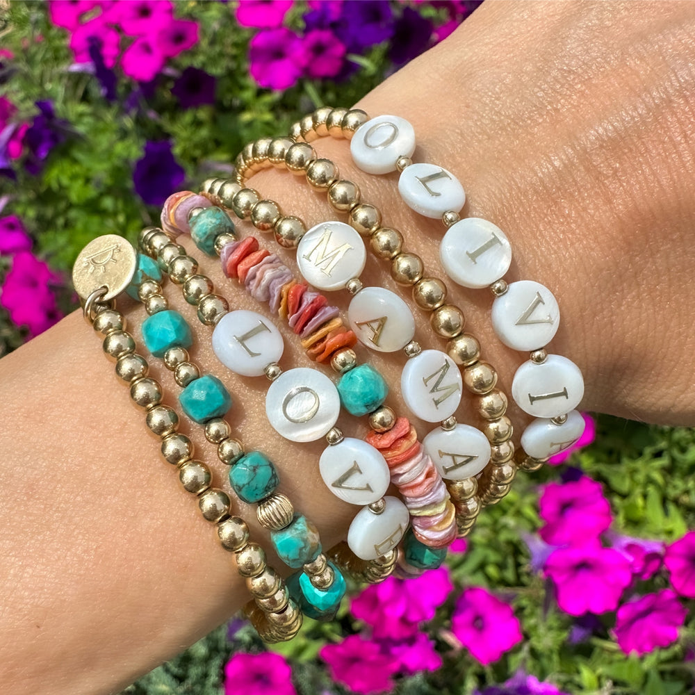 gold beaded bracelets with gemstones and personalized white alphabet beads on models wrist with flowers in background