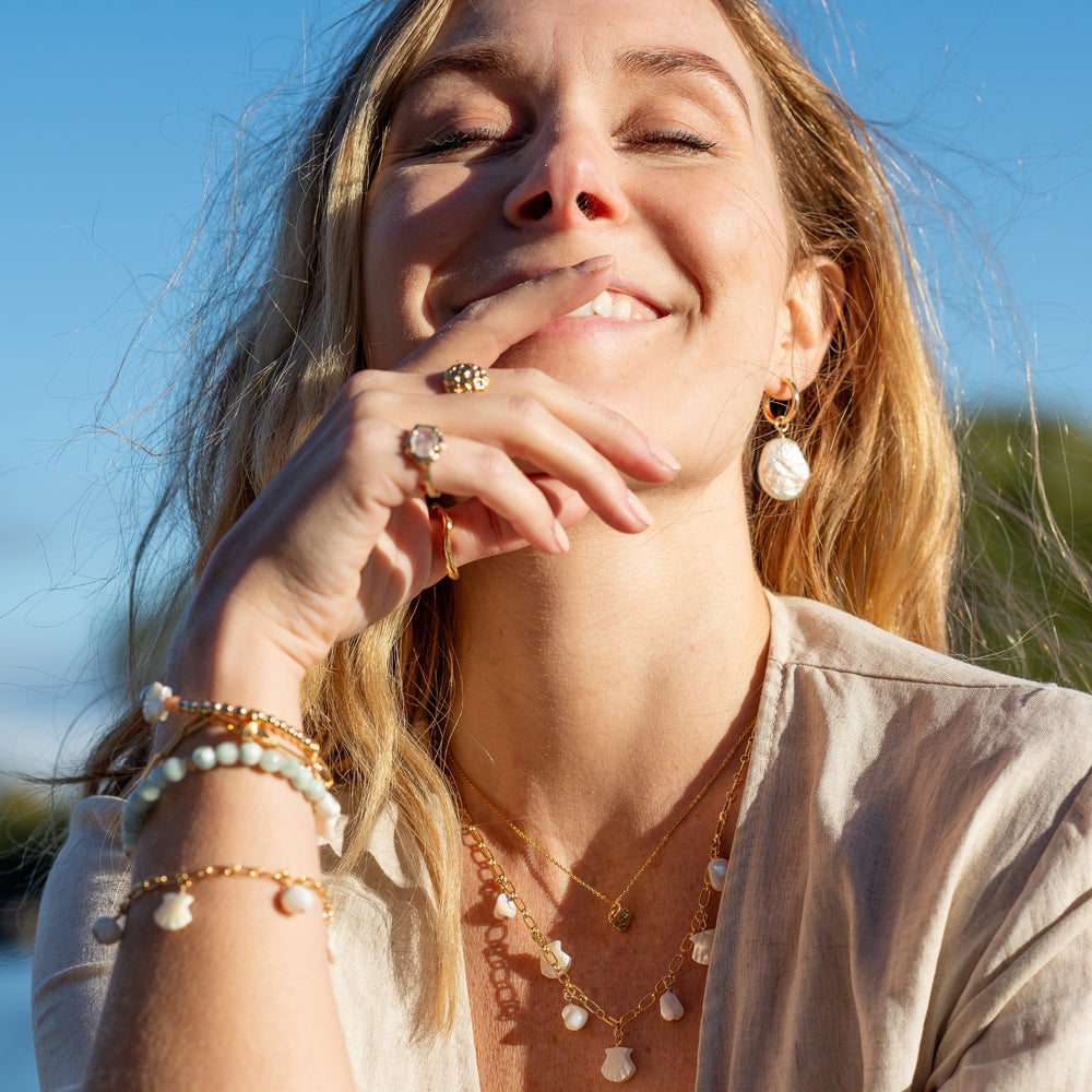 Close-up of a woman smiling with her eyes closed, wearing Ocean Whispers Shell Necklace and Ocean Mist Pearl Hoop Earrings by Blooming Lotus Jewelry. The  pearl charms add a natural elegance, perfectly complementing her layered jewelry look. The sunlight enhances the soft, beach-inspired aesthetic of her jewelry, exuding joy and carefree style.