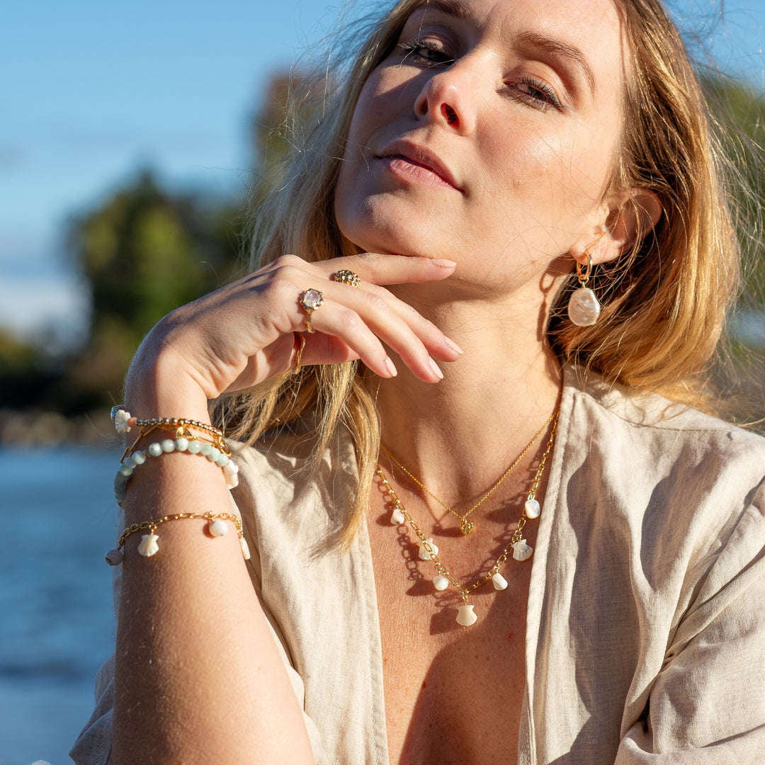 A woman wearing the Ocean Whispers necklace with mother-of-pearl shell charms, styled with layered gold necklaces, matching bracelets, a statement ring, and pearl drop earrings, set against a serene outdoor backdrop near water.