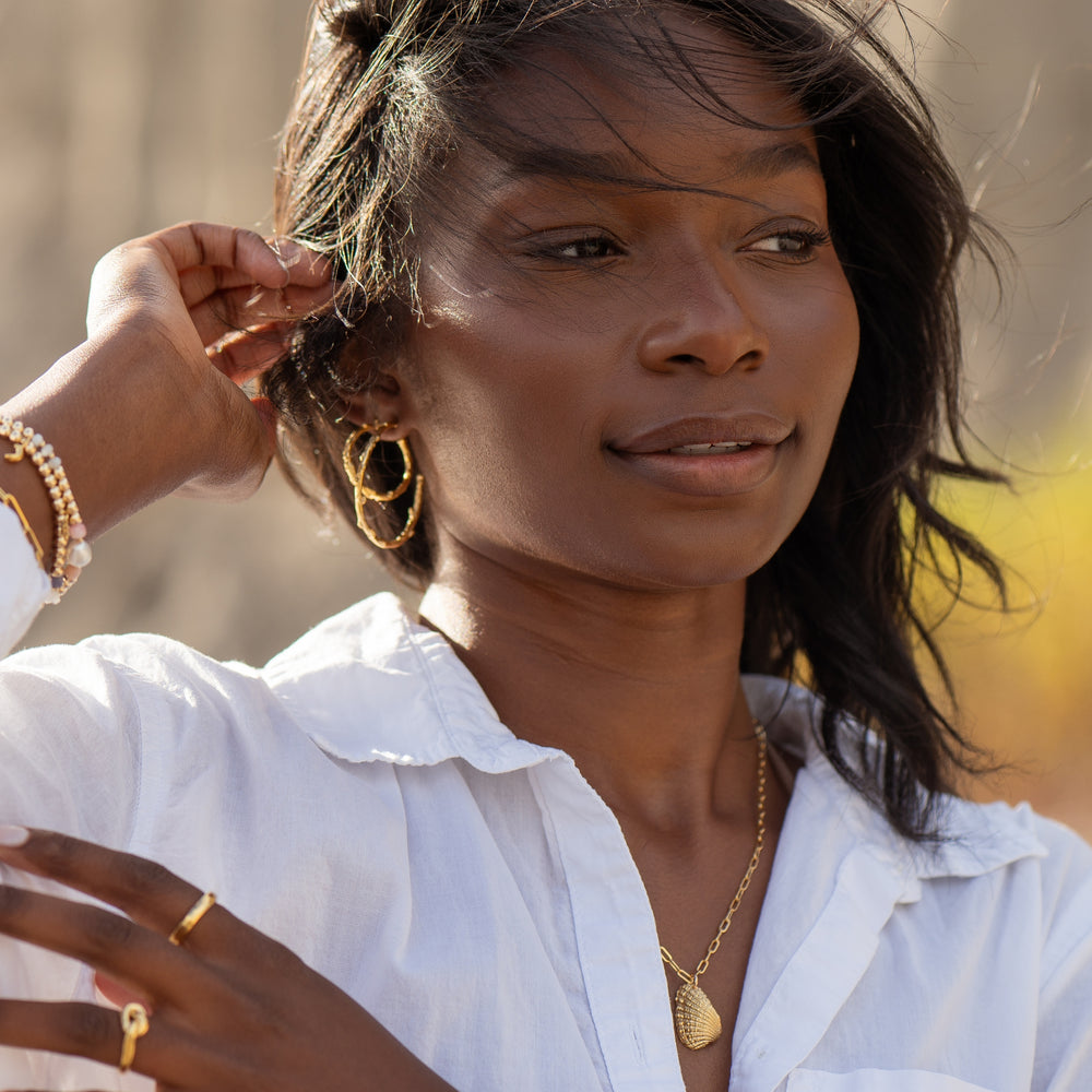 Woman wearing Large and Medium size Olive Branch Hoop Earrings in gold by Blooming Lotus Jewelry, paired with a gold shell pendant necklace and layered bracelets. The earrings feature a nature-inspired, textured branch design, adding a touch of boho elegance to her look.