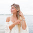 Woman by the water wearing large gold Olive Branch hoop earrings with a nature-inspired design, paired with a boho lace top, layered necklaces, stacked bracelets, and multiple rings. Her look is effortless and beachy, embodying a peaceful, free-spirited style