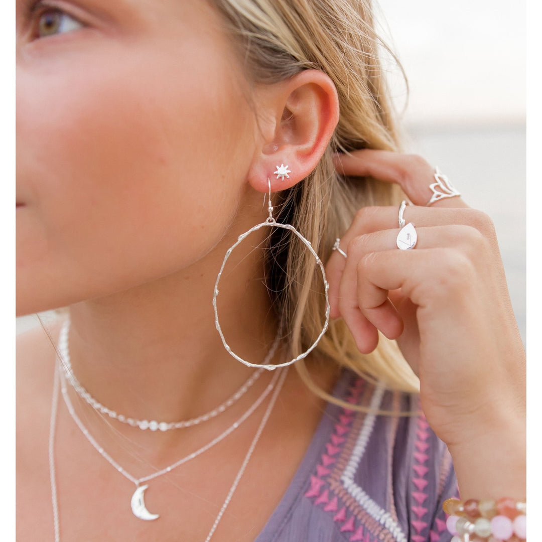 Close-up of a woman wearing large, silver Olive Branch hoop earrings in an organic, looped shape, symbolizing peace and resilience. She is also adorned with layered necklaces, a mix of rings, and bracelets, adding a boho-chic touch to her beach-inspired look