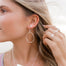 Close-up of woman wearing medium silver Olive Branch hoop earrings, heart stud earring, and multiple rings. She is holding some strands of her hair giving a relaxed boho style.