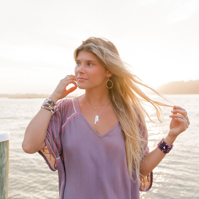 Woman at sunset wearing medium silver Olive Branch hoop earrings, layered bar necklaces, and stacked beaded bracelets. Her hair flows in the breeze, complementing her relaxed boho style with a light lavender top. The serene setting and natural jewelry capture a peaceful, free-spirited vibe.