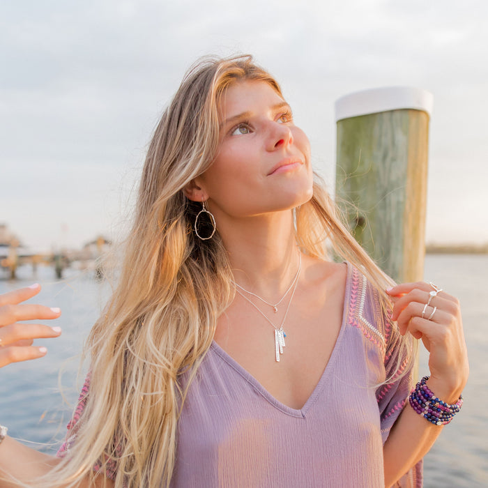 Woman at sunset wearing medium silver Olive Branch hoop earrings, layered bar necklaces, and stacked beaded bracelets. She is holding some strands of her hair giving a relaxed boho style with a light lavender top. The serene setting and natural jewelry capture a peaceful, free-spirited vibe.