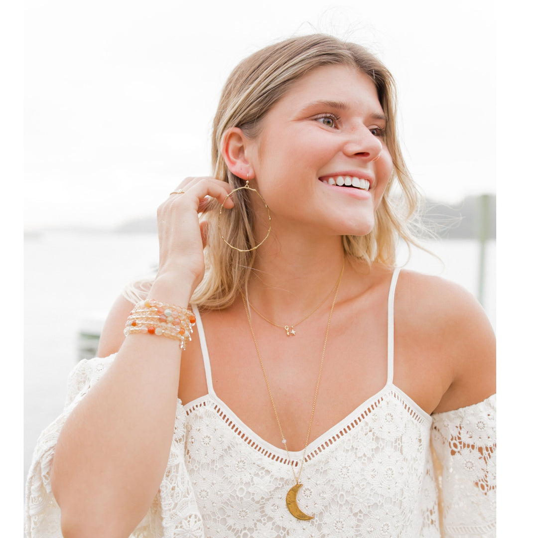 Close-up of woman by the water wearing large gold Olive Branch hoop earrings with a nature-inspired design, paired with a boho lace top, layered necklaces, stacked bracelets, and multiple rings. Her look is effortless and beachy, embodying a peaceful, free-spirited style