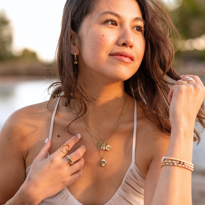 A woman outdoors wearing layered gold necklaces with organic pendants, gold earrings, and an open heart ring, paired with a gold band and beaded bracelets, radiating a natural, elegant, and boho-inspired style.