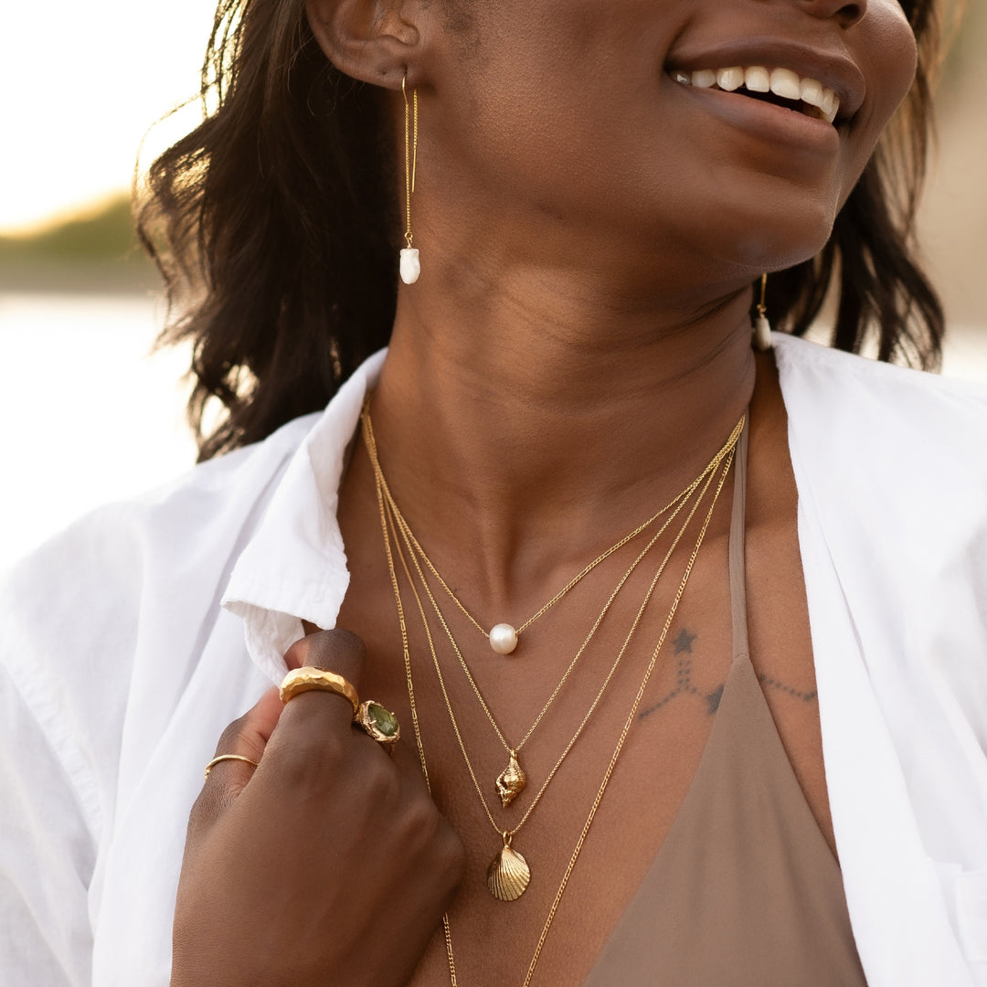 Close-up of a woman wearing layered gold necklaces, including the Pearl Drift Necklace with a single pearl, alongside shell-inspired pendants. She pairs them with a white shirt, shell drop earrings, and gold rings, exuding a natural, beachy elegance against a soft outdoor background.