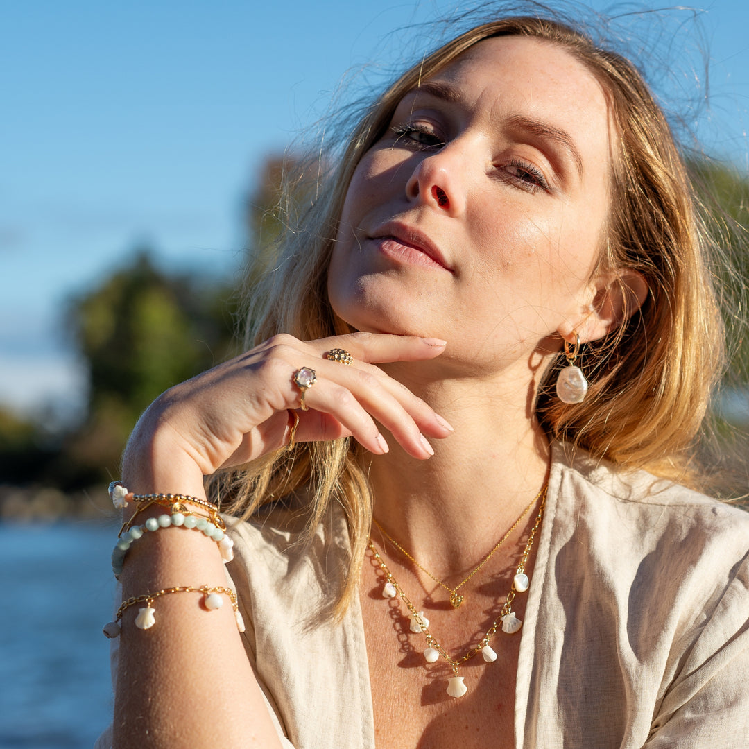 Woman wearing Ocean Mist Pearl Hoop Earrings by Blooming Lotus Jewelry. The large, organic freshwater pearl charms dangle elegantly from gold hoops, adding a touch of seaside sophistication to her look. She pairs the earrings with layered shell necklaces and bracelets, creating a cohesive, beach-inspired ensemble that glows in the natural sunlight.