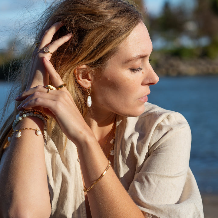 Side profile of a woman wearing Ocean Mist Pearl Hoop Earrings by Blooming Lotus Jewelry. The elongated freshwater pearl charm hangs gracefully from a gold hoop, adding a touch of coastal elegance to her look. Her layered bracelets and rings, paired with a soft beige top, create a natural, beach-inspired aesthetic against the serene water backdrop.