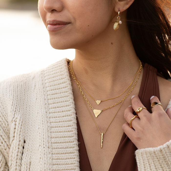 Close-up of a woman wearing layered gold necklaces, including the Soul on Fire Necklace with a sleek spear-shaped pendant, paired with geometric pendants and a cozy cream sweater. She also wears gold rings and delicate drop earrings, creating a warm, modern boho look.