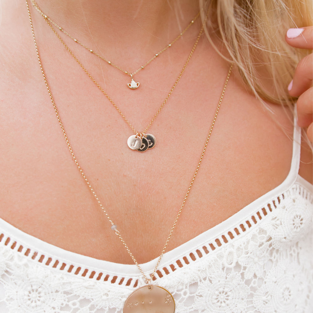 Close-up of layered gold necklaces on a woman wearing a white lace top. The top layer features a tiny lotus charm, the middle layer showcases small initial pendants, and the longest layer features a large round pendant engraved with celestial designs for a delicate and personalized look.