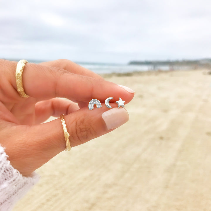 Close-up of a hand holding three tiny silver stud earrings in the shapes of a rainbow, crescent moon, and star, with a beach and cloudy sky in the background. The hand also wears textured branch gold rings, adding a natural and minimalist aesthetic."