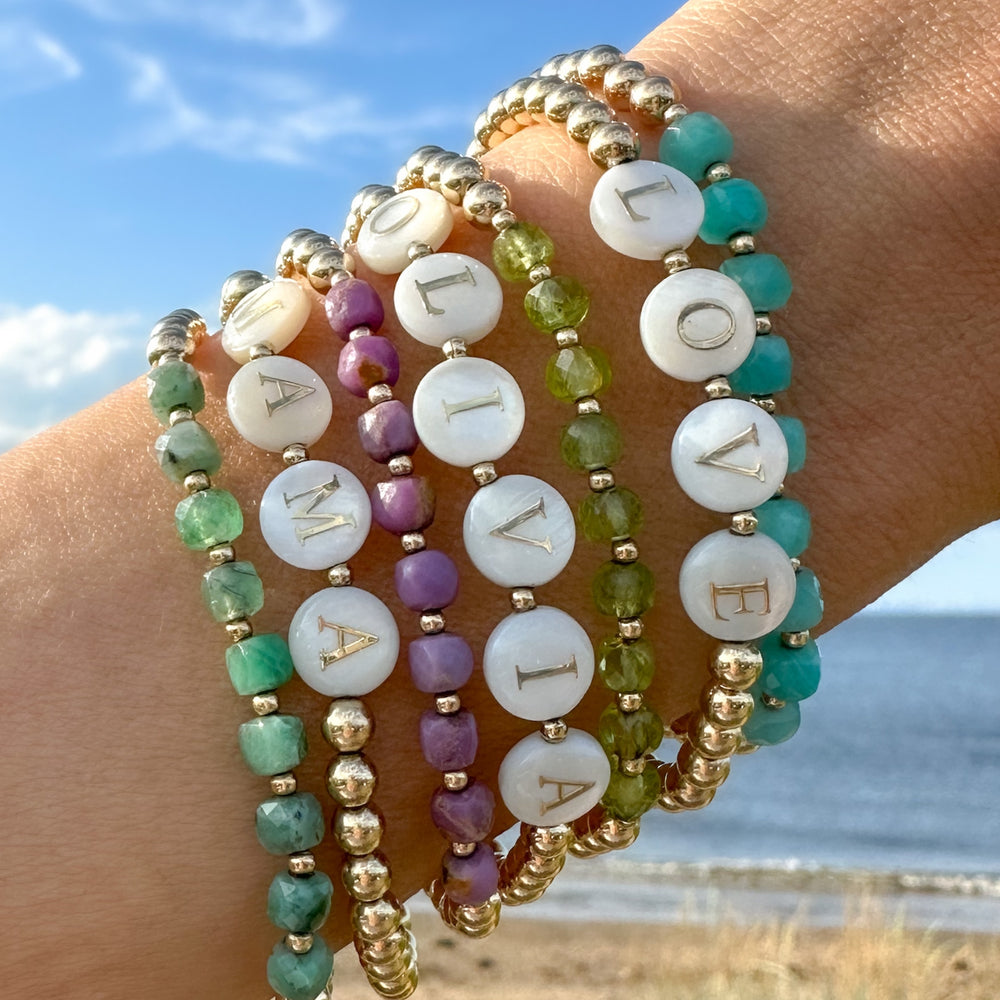 Stack of gold beaded bracelets with mother-of-pearl letter beads spelling "LOVE," "MAMA," and "OLIVIA," accented with faceted colorful gemstones in shades of green, purple, and turquoise. Worn on a wrist against a beach backdrop with blue skies and ocean waves. By Blooming Lotus Jewelry.