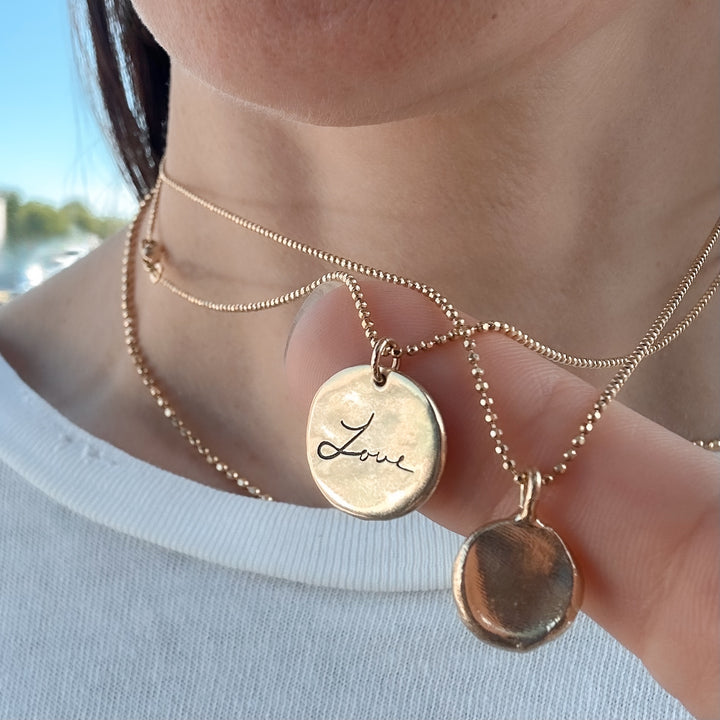 Close-up of a woman wearing two gold necklaces: one featuring the 'Written With Love' pendant engraved with custom handwriting reading 'Love,' and the other with a fingerprint pendant, both suspended on delicate gold chains – Blooming Lotus Jewelry.