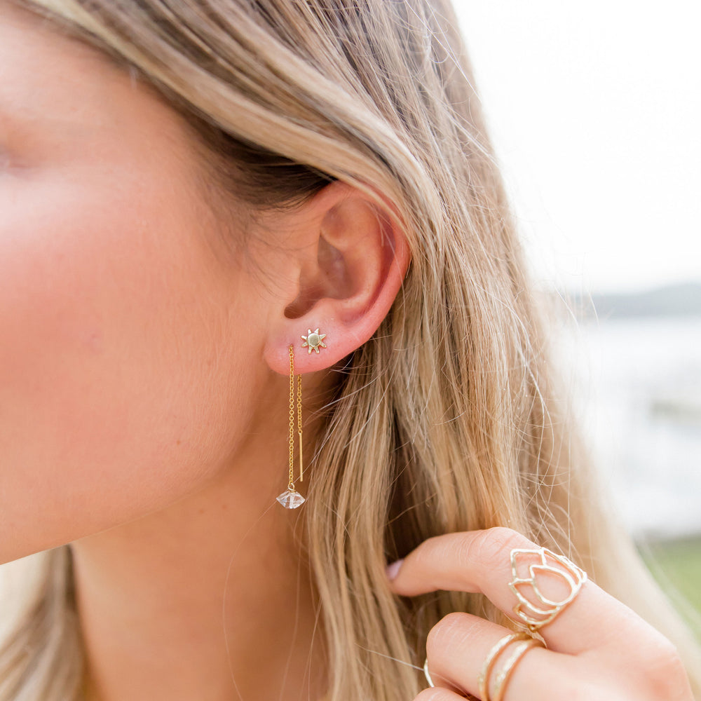 A close-up of a woman's ear wearing a 14k gold Soleil Sun Stud earring paired with a delicate gold threader earring featuring a faceted Herkimer Diamond crystal drop. Her blonde hair is tucked behind her ear, and she is also wearing gold stackable rings, including a lotus ring. The look is elegant and radiant, perfect for a light, boho-chic vibe. By Blooming Lotus Jewelry