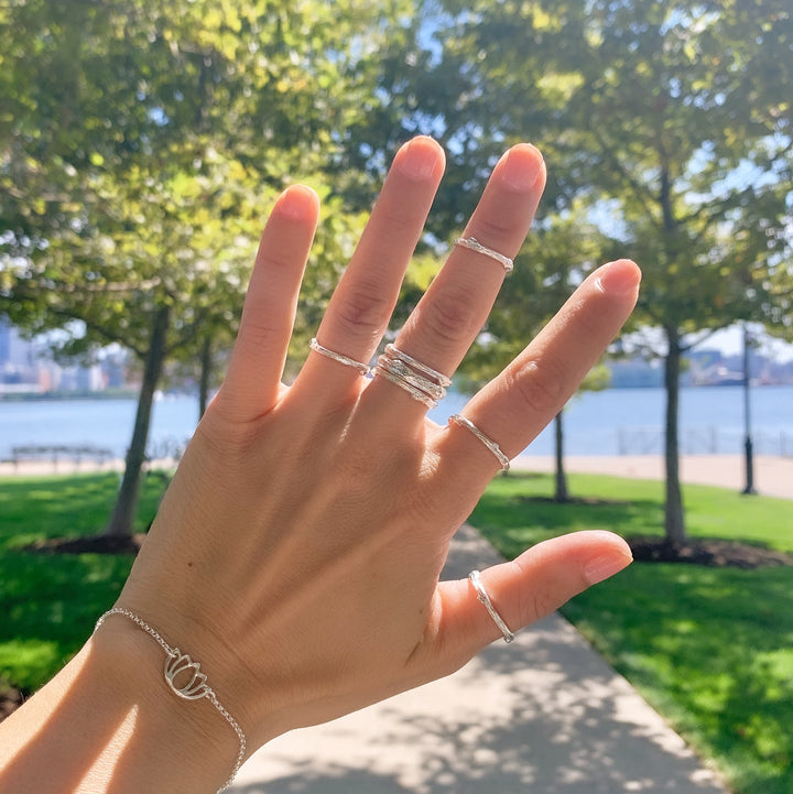 Hand adorned with silver Olive Branch twig-inspired stacking rings and a lotus charm bracelet, displayed outdoors on a sunny day with a scenic park and waterfront in the background, creating a natural and serene vibe.