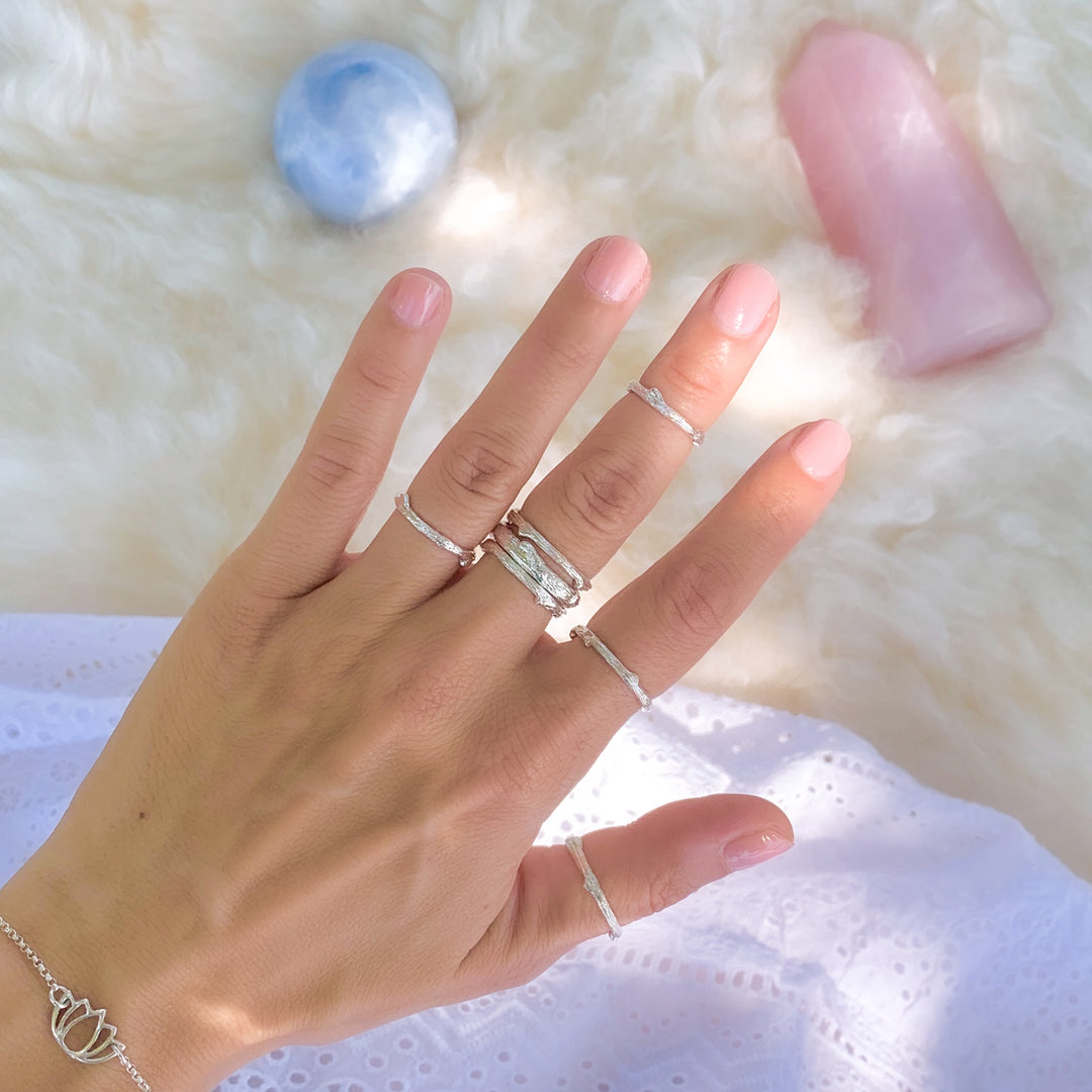 Hand showcasing a collection of silver twig-inspired stacking rings, delicately styled on each finger, paired with a lotus charm bracelet. The background features a soft, fluffy surface adorned with a rose quartz tower and a blue gemstone sphere, exuding a serene and natural vibe.