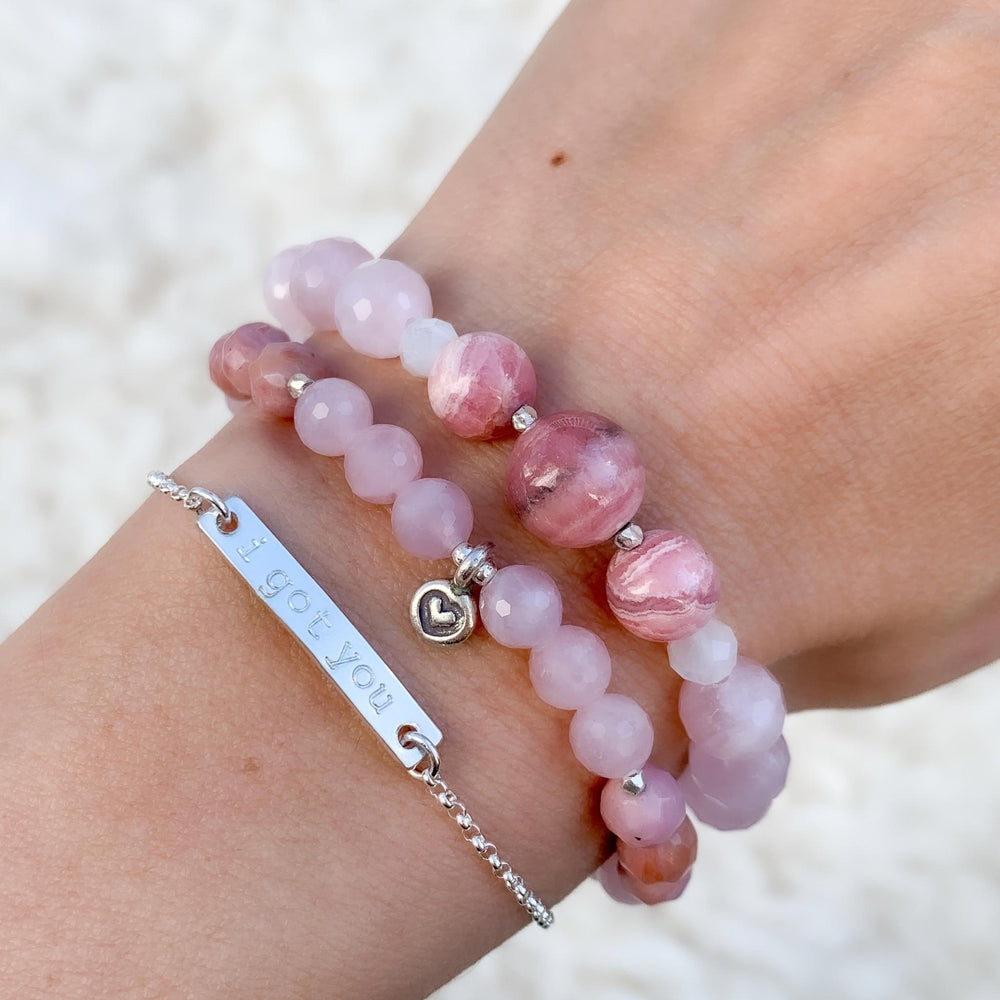 Close-up image of a wrist adorned with a stack of bracelets, including the 'Choose Love' bracelet featuring rose quartz and rhodonite gemstones with a tiny silver heart charm. Paired with a minimalist silver bar bracelet hand-stamped with 'I got you,' this stylish combination radiates love, support, and positive energy, set against a soft, neutral background.