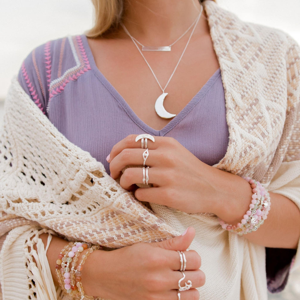 Close-up of a woman wearing layered celestial jewelry, including the Eye of Protection Evil Eye Ring, Luna Crescent Moon Ring, and other silver stacking rings. She is styled with a crescent moon necklace, delicate bracelets with pink gemstones, and a cozy boho knit shawl over a lavender top, creating a dreamy and boho look.