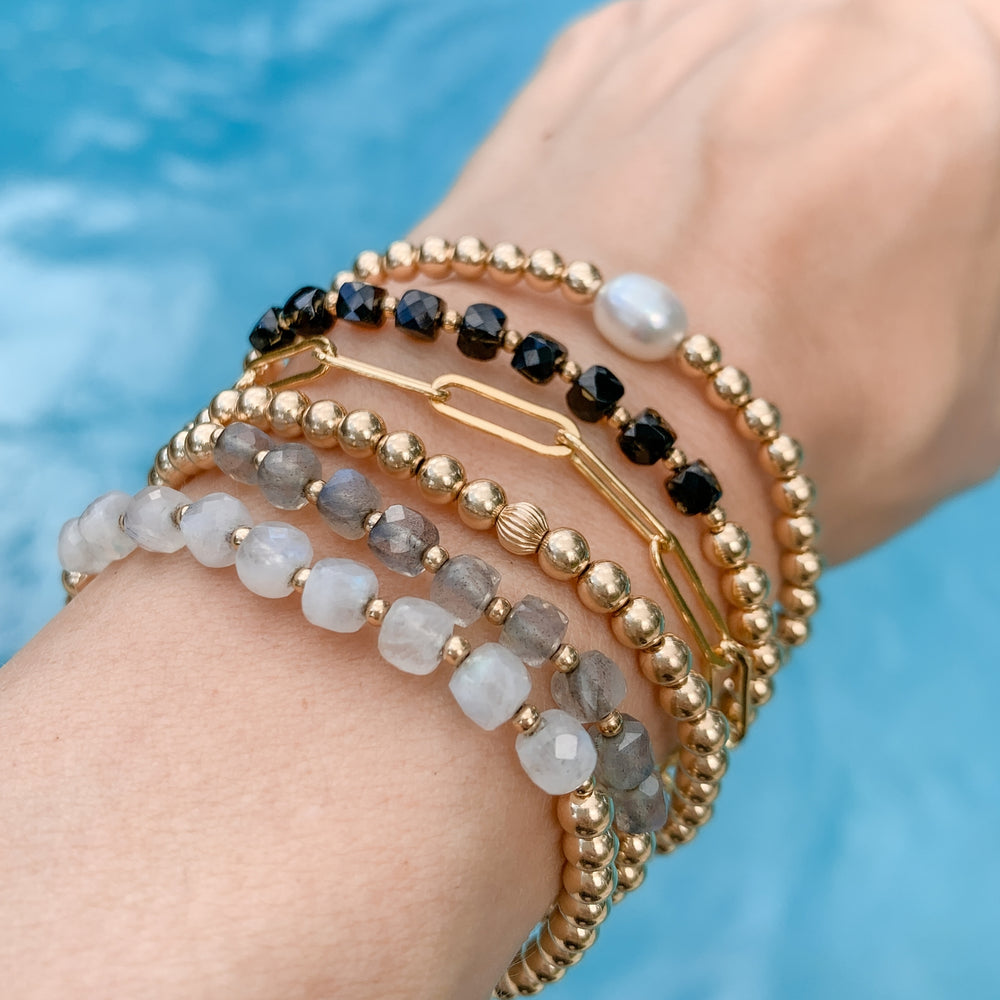 Stack of gold beaded bracelets featuring faceted moonstone, labradorite, black spinel, a freshwater pearl, and a gold paperclip chain. Worn on a wrist against a blue water background, creating an coastal-inspired and boho layered look.