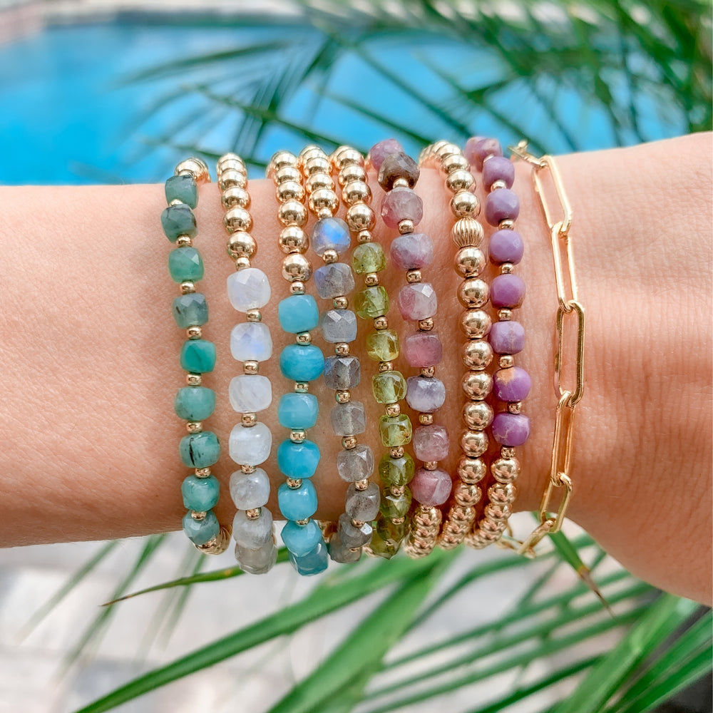 Stack of gold beaded bracelets featuring faceted emerald, moonstone, amazonite,  labradorite, peridot, phosphosiderite, and a gold paperclip chain. Worn on a wrist against a bright blue water background, creating a coastal inspired and boho layered look.
