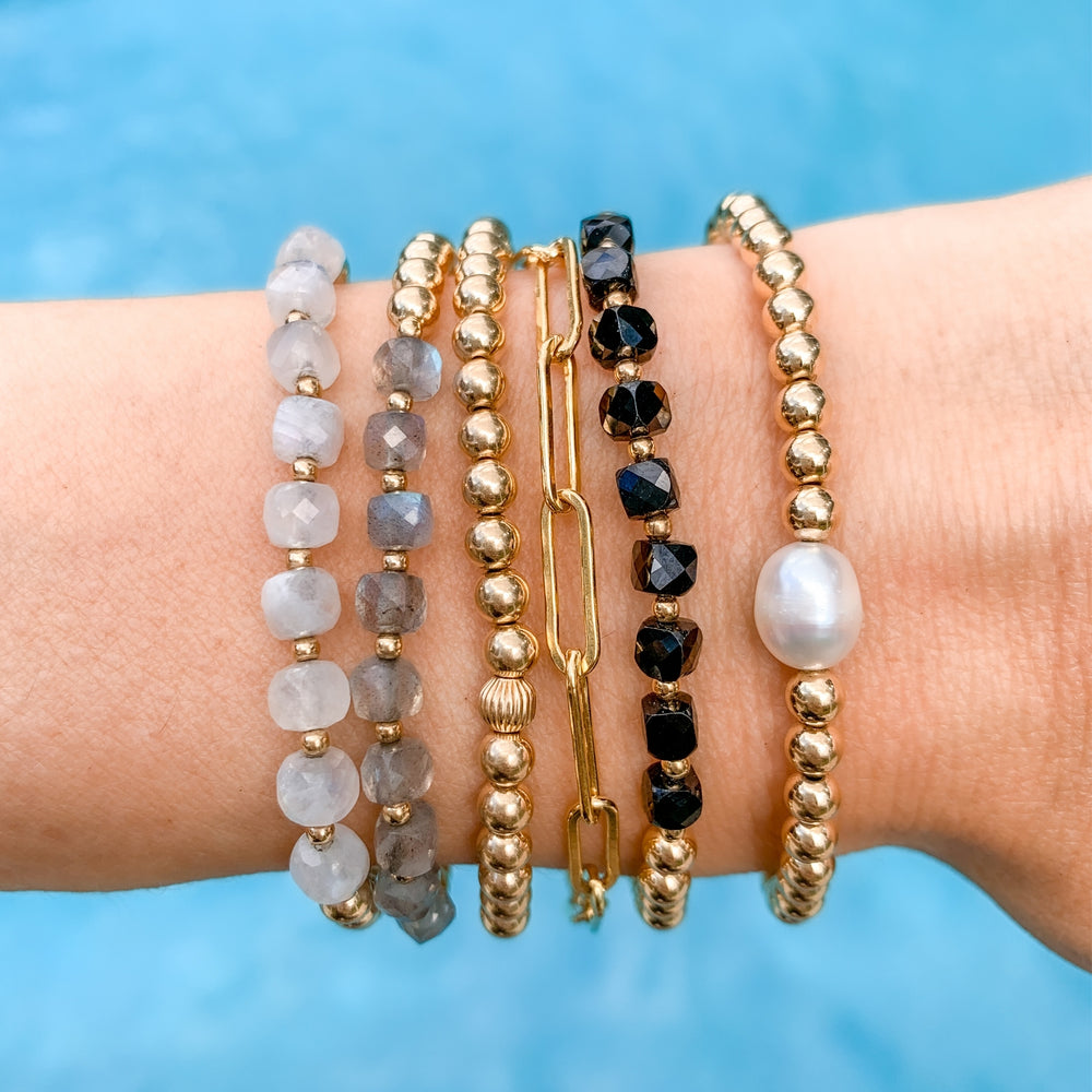 Stack of gold beaded bracelets featuring faceted moonstone, labradorite, black spinel, a freshwater pearl, and a gold paperclip chain. Worn on a wrist against a bright blue water background, creating a coastal inspired and boho layered look.