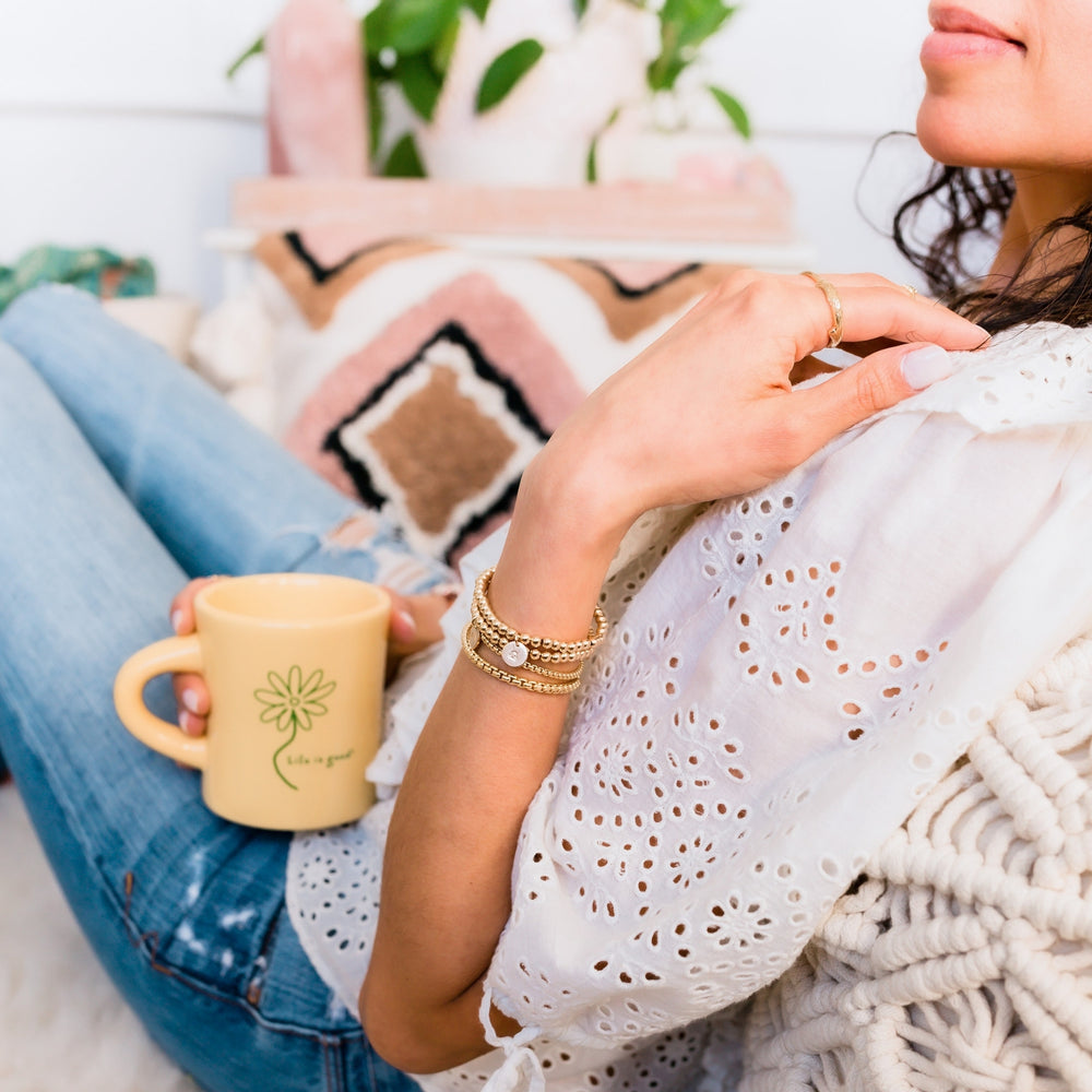 Close-up of a woman relaxing in a cozy, boho-inspired setting, wearing layered gold beaded bracelets with personalized charms. She holds a yellow mug with 'Life is Good' printed on it, while dressed in a delicate white eyelet blouse, complemented by macrame decor and warm, earthy tones in the background.