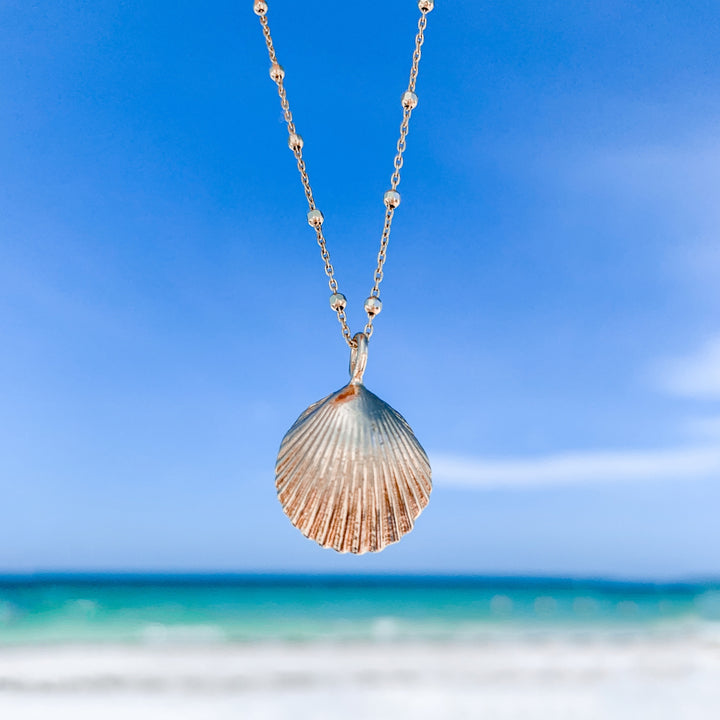 gold shell pendant hanging on gold bead chain with beach ocean in background - blooming lotus jewelry