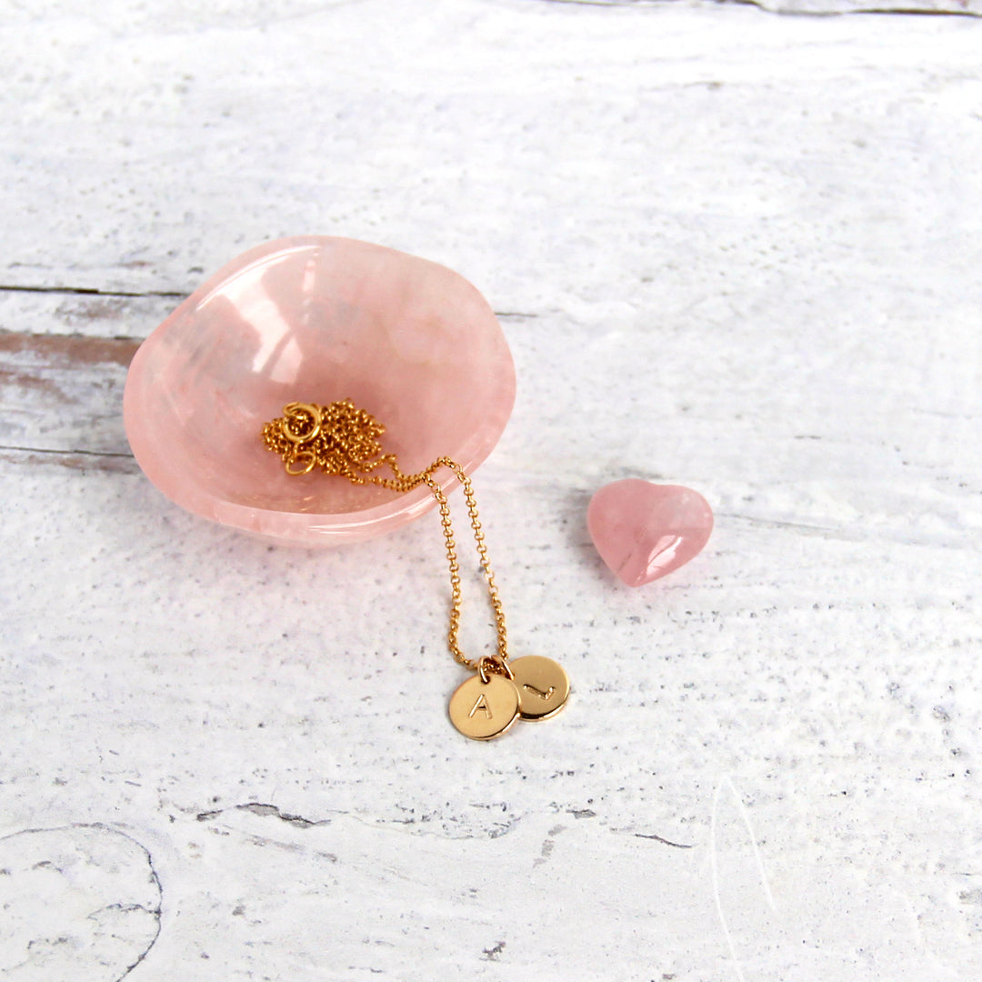 two Gold Tiny Initial Disc Coin necklaces on gold chain in rose quartz dish